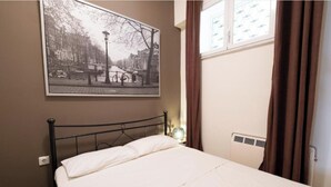A bedroom with a bed, a wall-mounted picture of a city street, a window with curtains, and a radiator. - Levanda Syros with Free Parking (Ermoupoli)