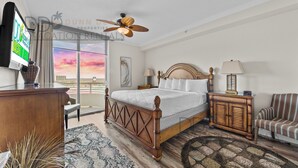A hotel room with a bed, a ceiling fan, a TV, a desk, a chair, and a view of the sunset. - Ocean Club 408 | Enjoy Views of the Gulf! (Biloxi)