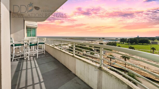 Un balcon donnant sur une plage, avec vue sur le coucher de soleil, une balustrade blanche et une table avec des chaises.