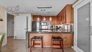 Une cuisine moderne dotée de plans de travail en granit, d’appareils électroménagers en acier inoxydable et de meubles en bois. On y trouve deux tabourets de bar et un réfrigérateur.
