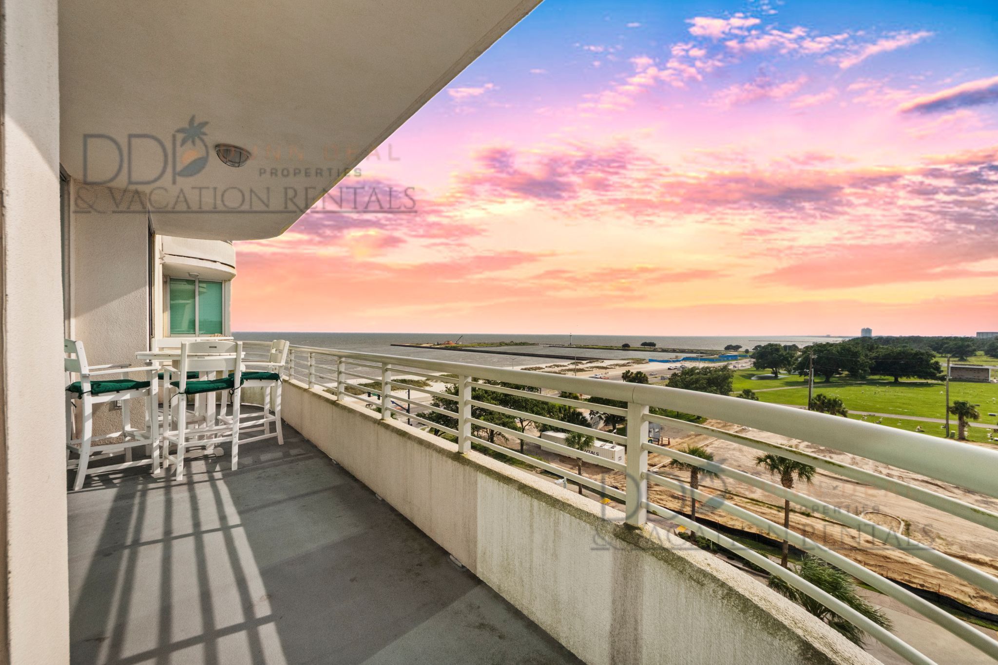 Un balcon donnant sur une plage, avec vue sur le coucher de soleil, une balustrade blanche et une table avec des chaises.