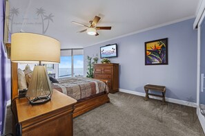 A bedroom with a bed, a nightstand, a desk, a chair, a ceiling fan, a TV, and a painting.
