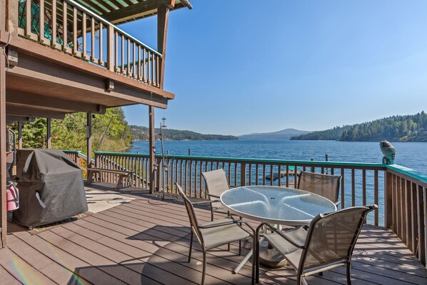 A wooden deck with a glass table and chairs overlooking a lake.