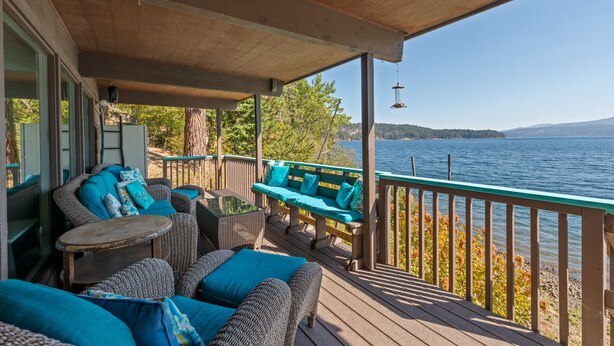 Une terrasse couverte, dotée de meubles en osier et de coussins turquoise, qui offre une vue sur un lac et est agrémentée d’une balustrade en bois.