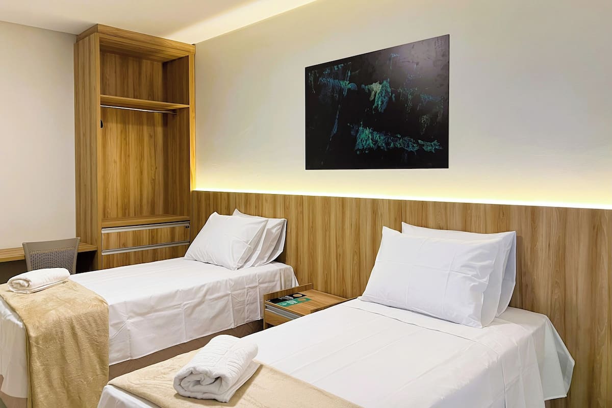 A hotel room with two beds, a wooden wardrobe, a wall-mounted artwork, and a small table with a magazine.