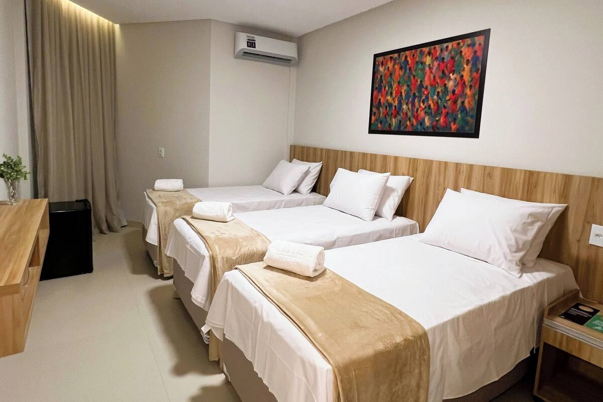 A hotel room with three single beds, a wooden headboard, a painting on the wall, and an air conditioning unit.