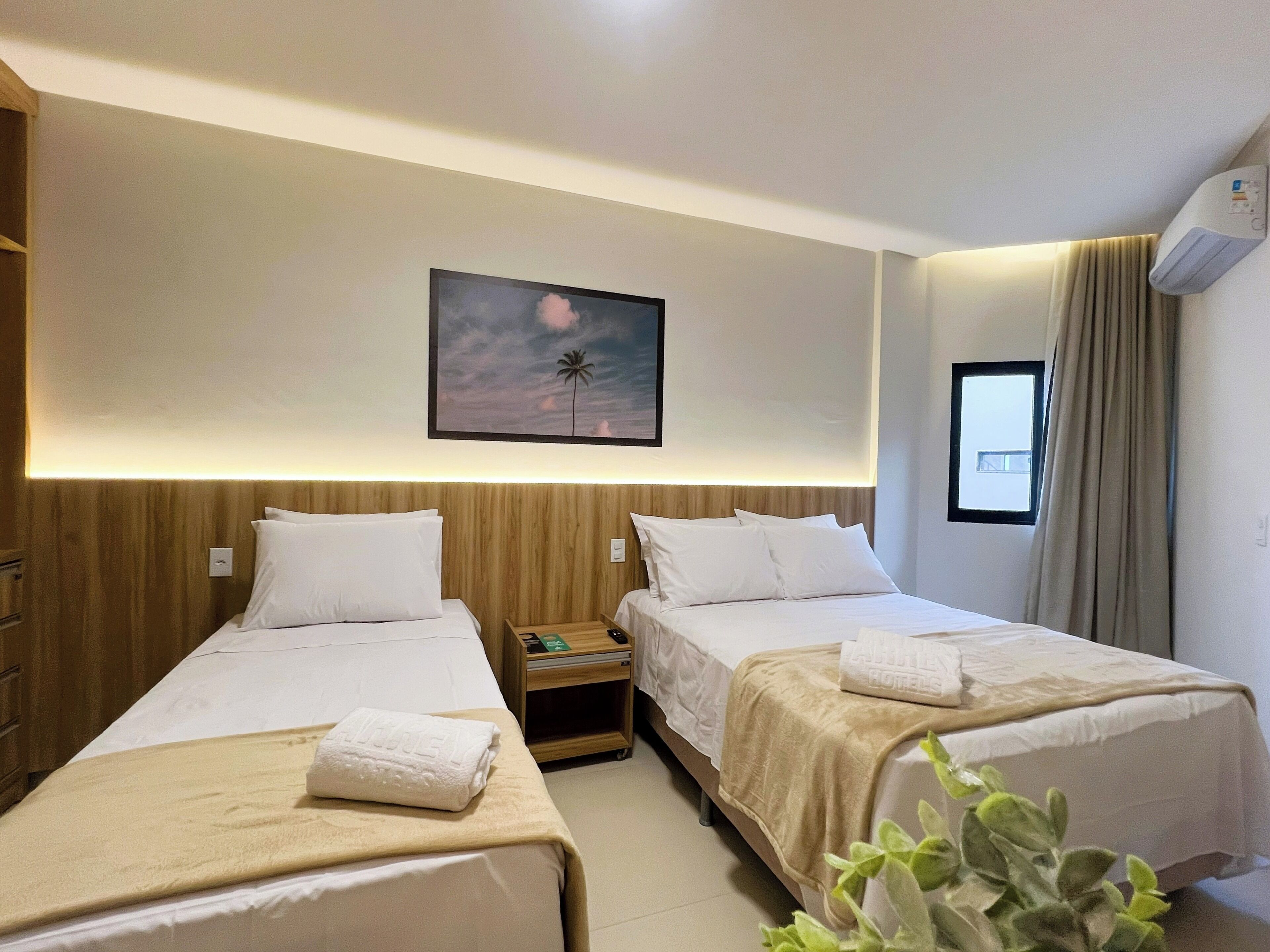 A hotel room with two single beds, a nightstand, a painting of a palm tree, a window with curtains, and an air conditioning unit.