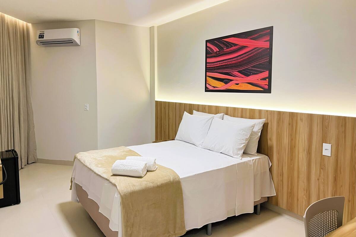 A hotel room with a bed, a wall-mounted air conditioner, a painting, and a chair.
