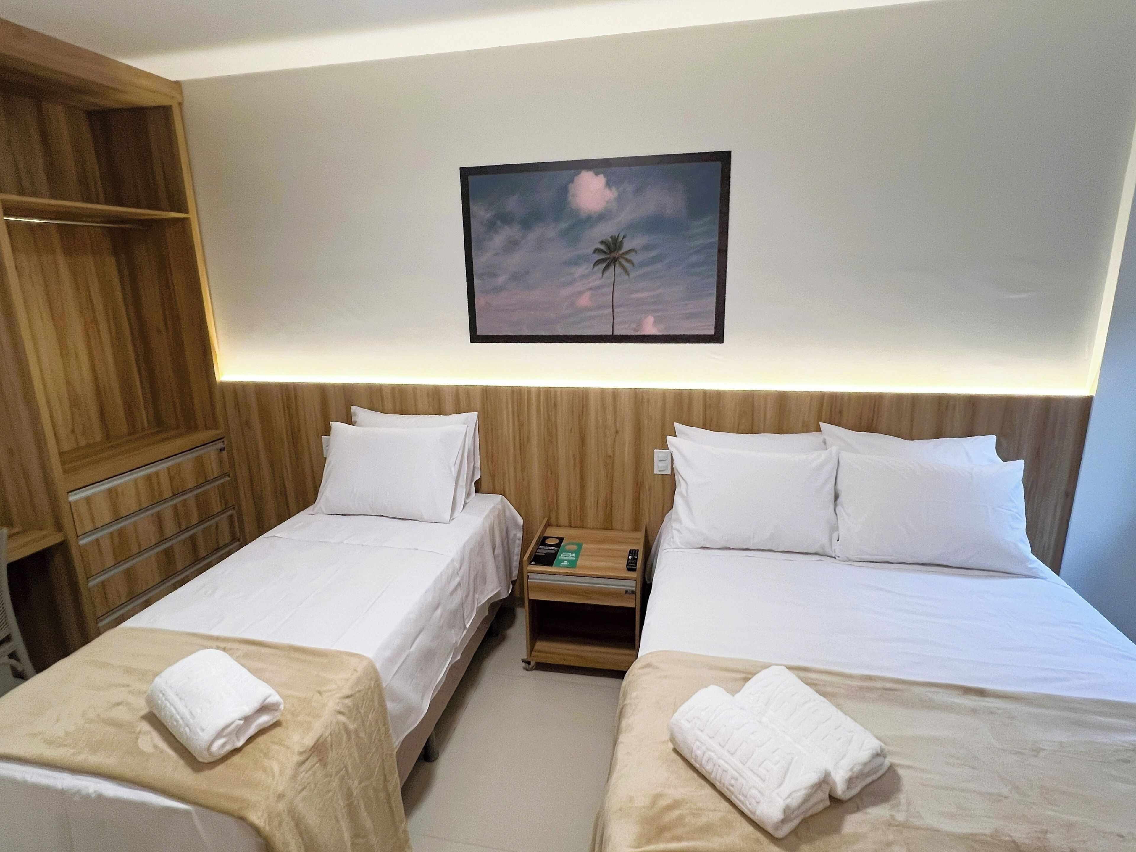 Ein Hotelzimmer mit zwei Einzelbetten, einem Holzschrank, einem Nachttisch mit Fernbedienung und einem gerahmten Bild an der Wand.