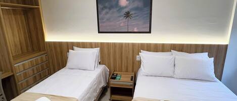 A hotel room with two single beds, a wooden wardrobe, a nightstand with a remote control, and a framed picture on the wall.