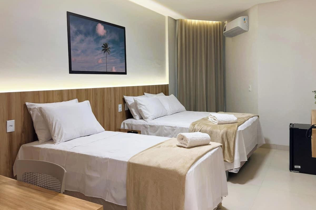 A hotel room with two beds, a wooden headboard, a painting of a palm tree, a window with curtains, and an air conditioning unit.