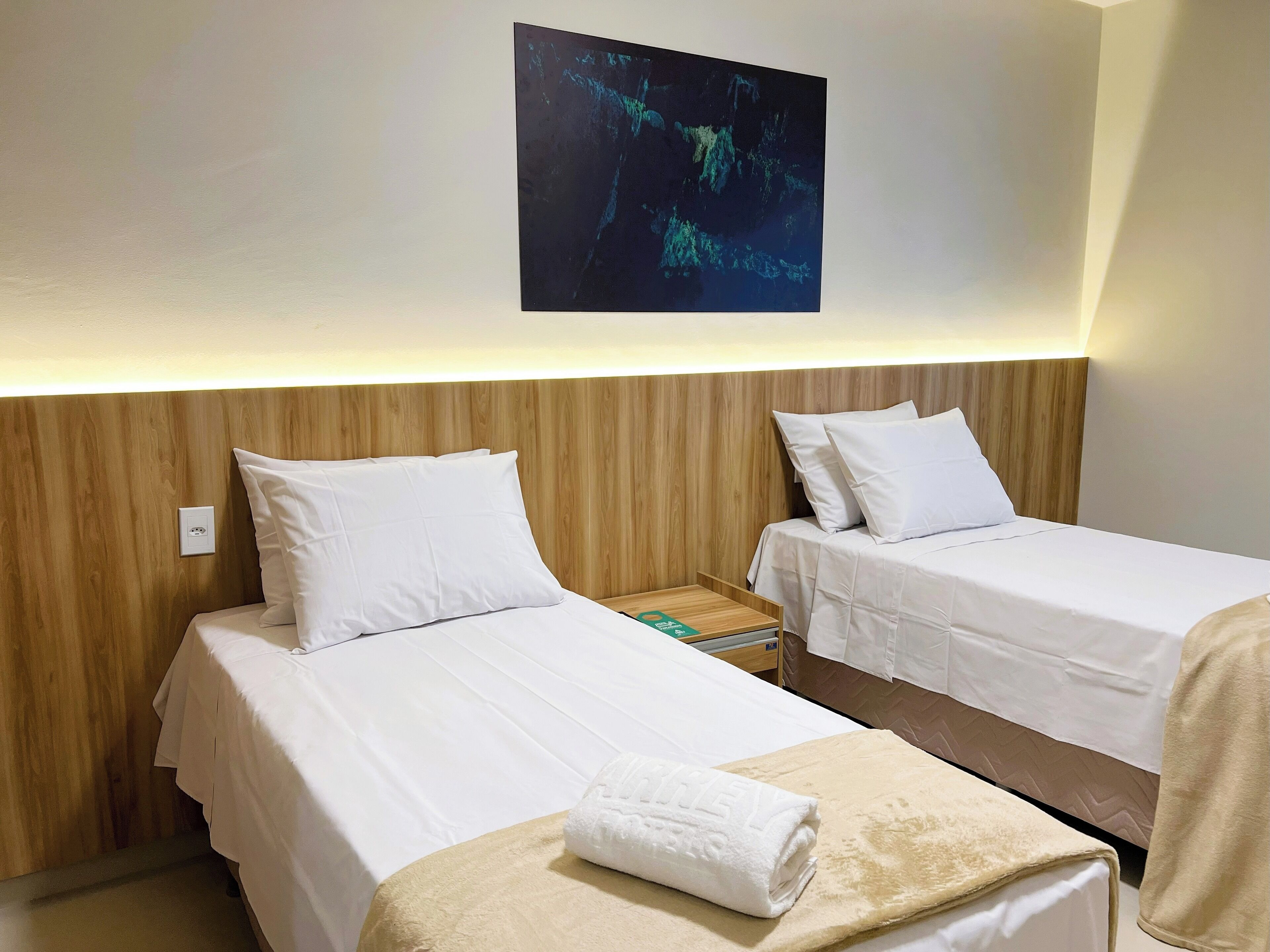 Quarto de hotel com duas camas de solteiro, cabeceira de madeira, mesa de cabeceira pequena e uma obra de arte na parede.