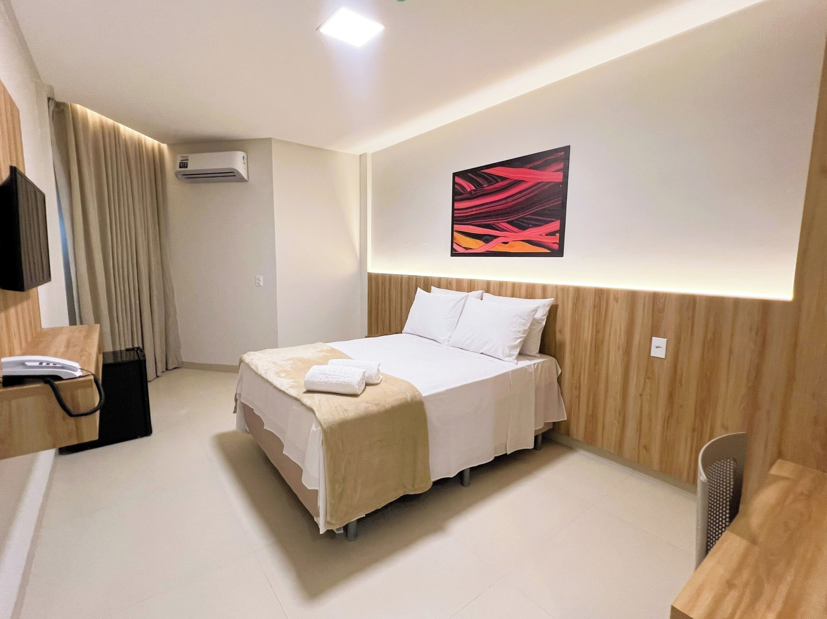 A hotel room with a bed, a desk, a chair, a TV, and an air conditioning unit.