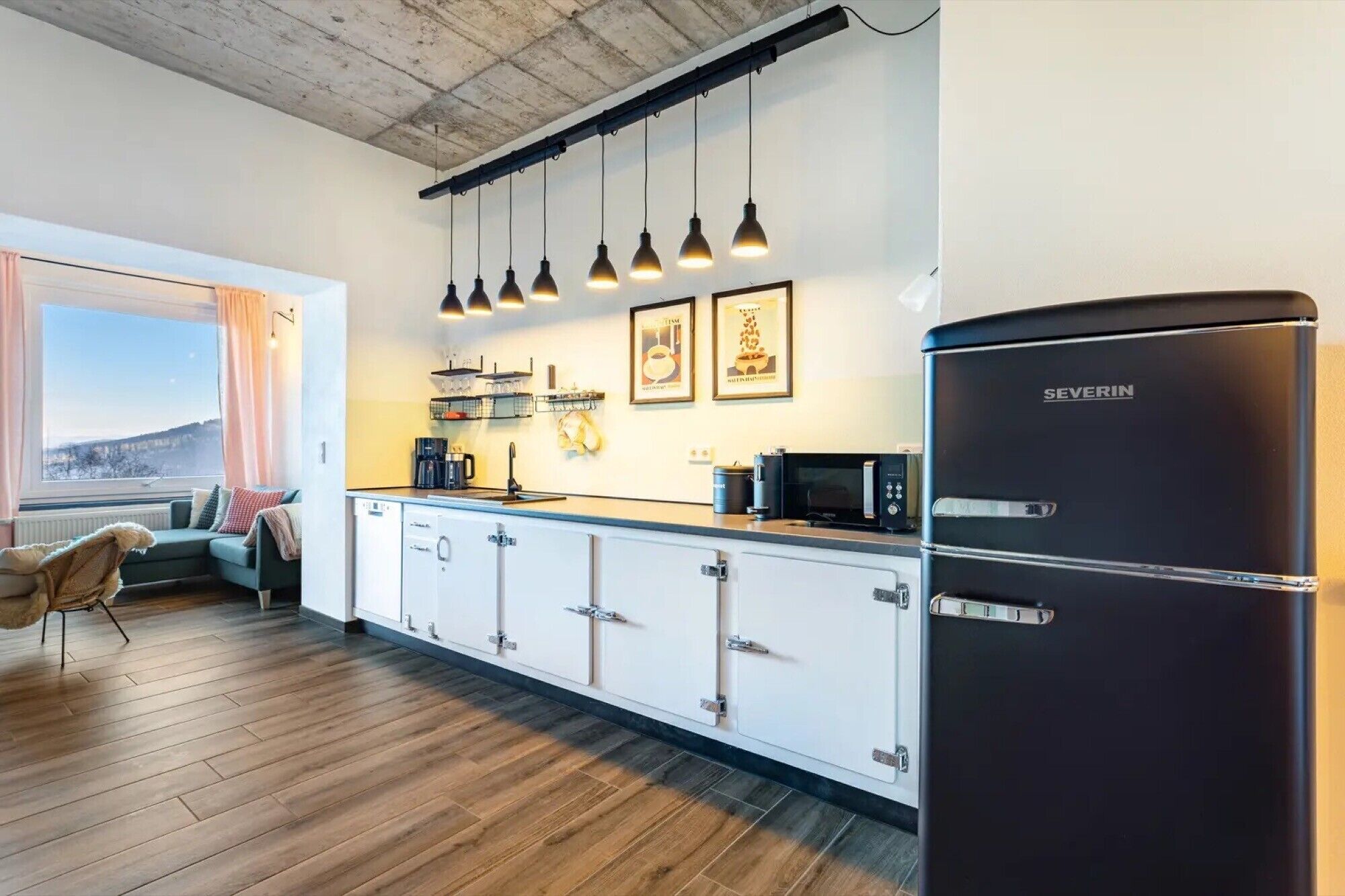A modern kitchen with a black refrigerator, wooden cabinets, a window with curtains, and hanging pendant lights.
