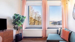 Uma sala de estar com sofá, televisão, planta em vaso e vista para árvores e colinas pelas janelas.