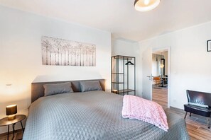 A bedroom with a bed, a bedside table, a hanging light, a coat rack, and a view into another room.