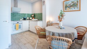 Een kleine keuken met witte kastjes, een groene spatwand en een eethoek met rieten stoelen en een ronde tafel.