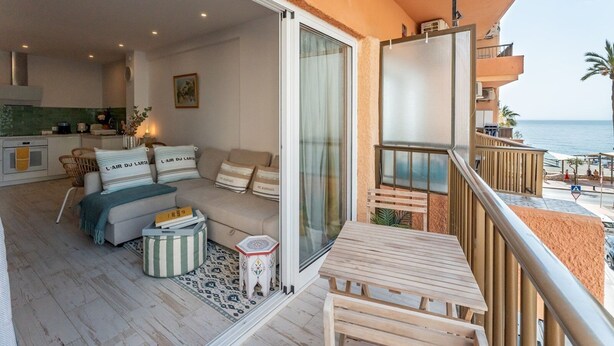 Een balkon met een houten tafel en stoelen, een bank met gestreepte kussens en uitzicht op zee.