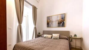 A bedroom with a large bed, a bedside table with a lamp, a wall-mounted abstract painting, and a window with curtains. - Casa Vico Ferruccio 24 (Noto (SR))