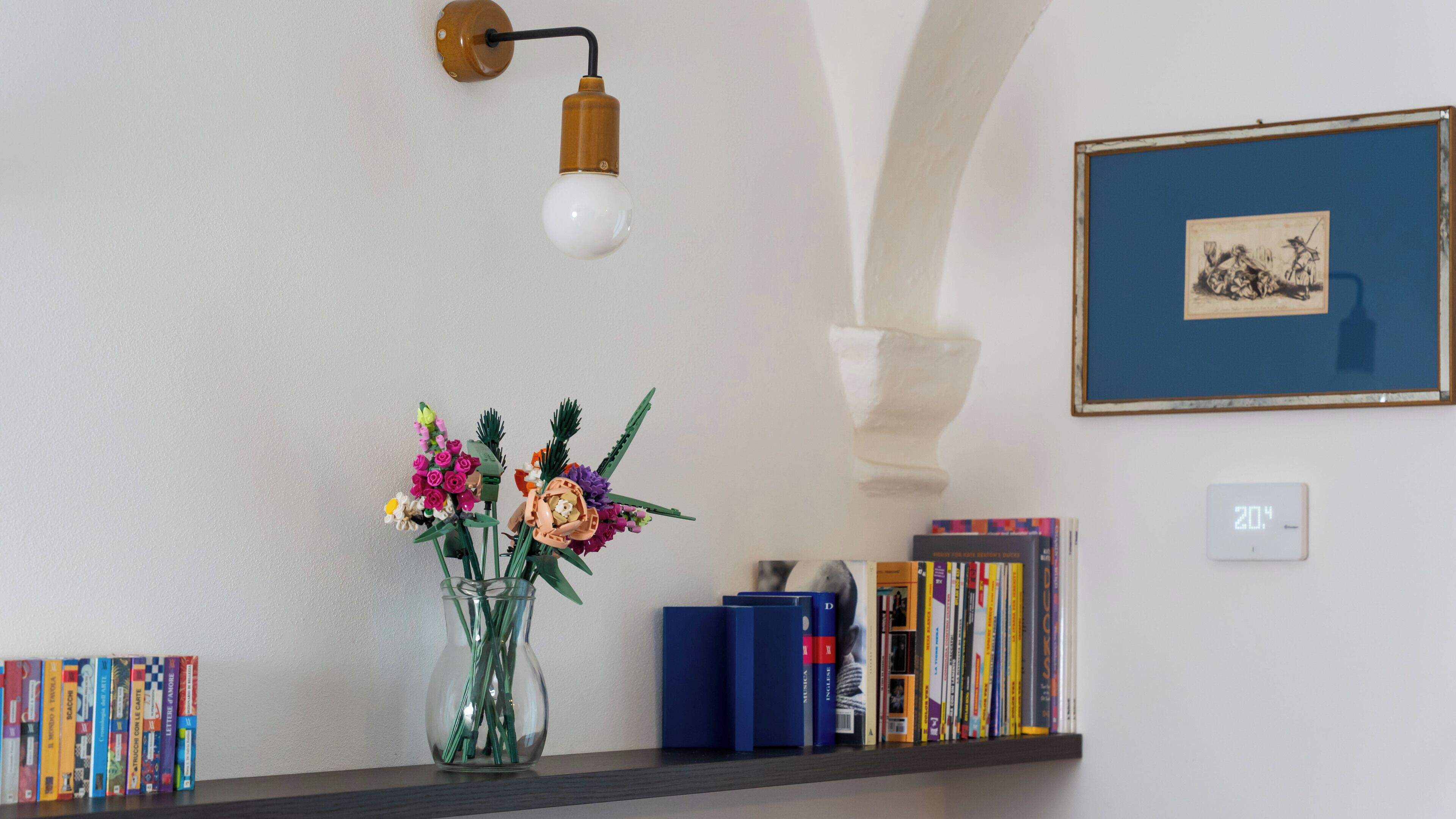 Una prestatgeria plena de llibres, un vas amb flors, una làmpada de paret i un quadre emmarcat.