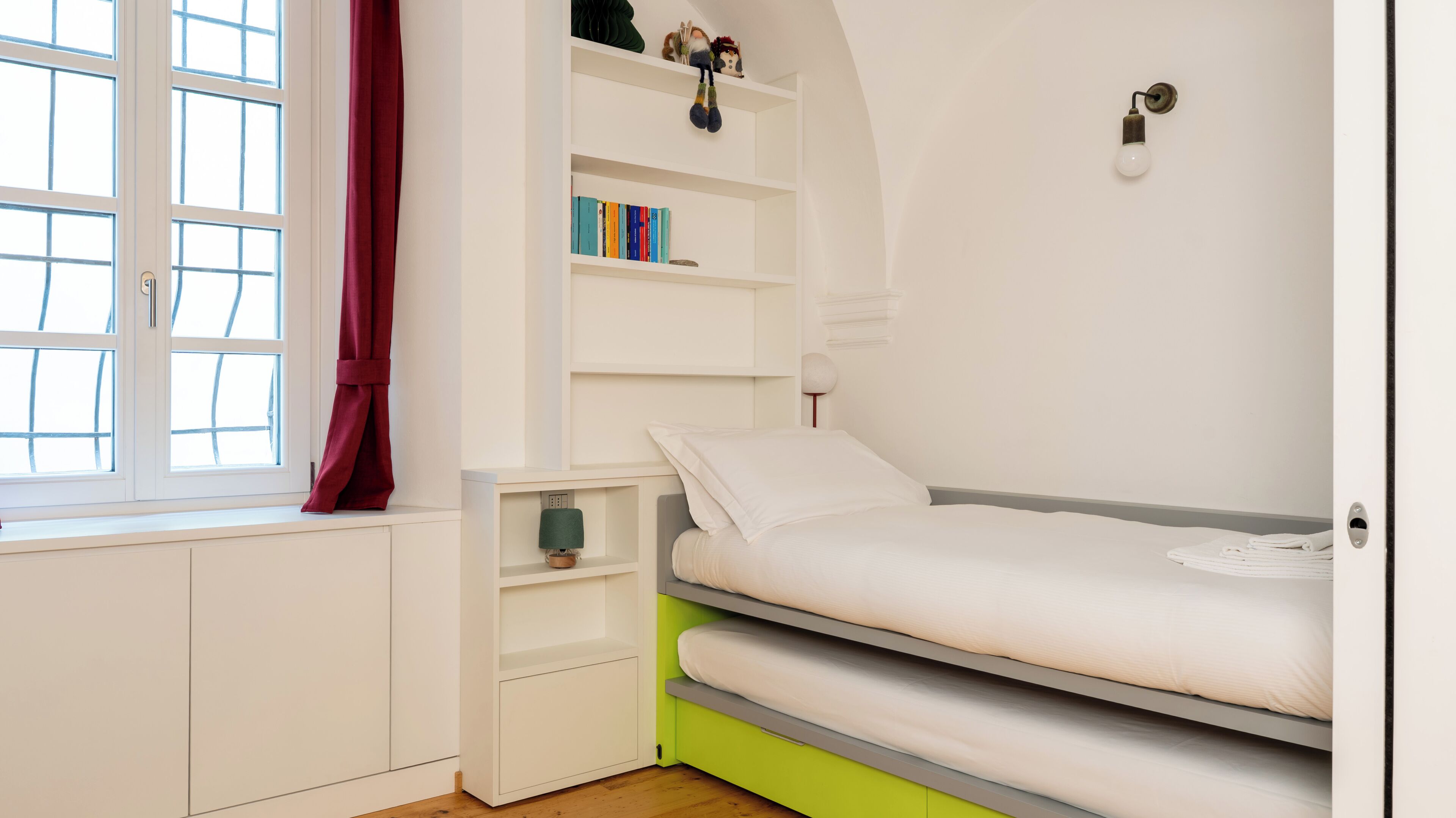 A bedroom with a bed, a window with curtains, a bookshelf, and a wall-mounted light fixture.