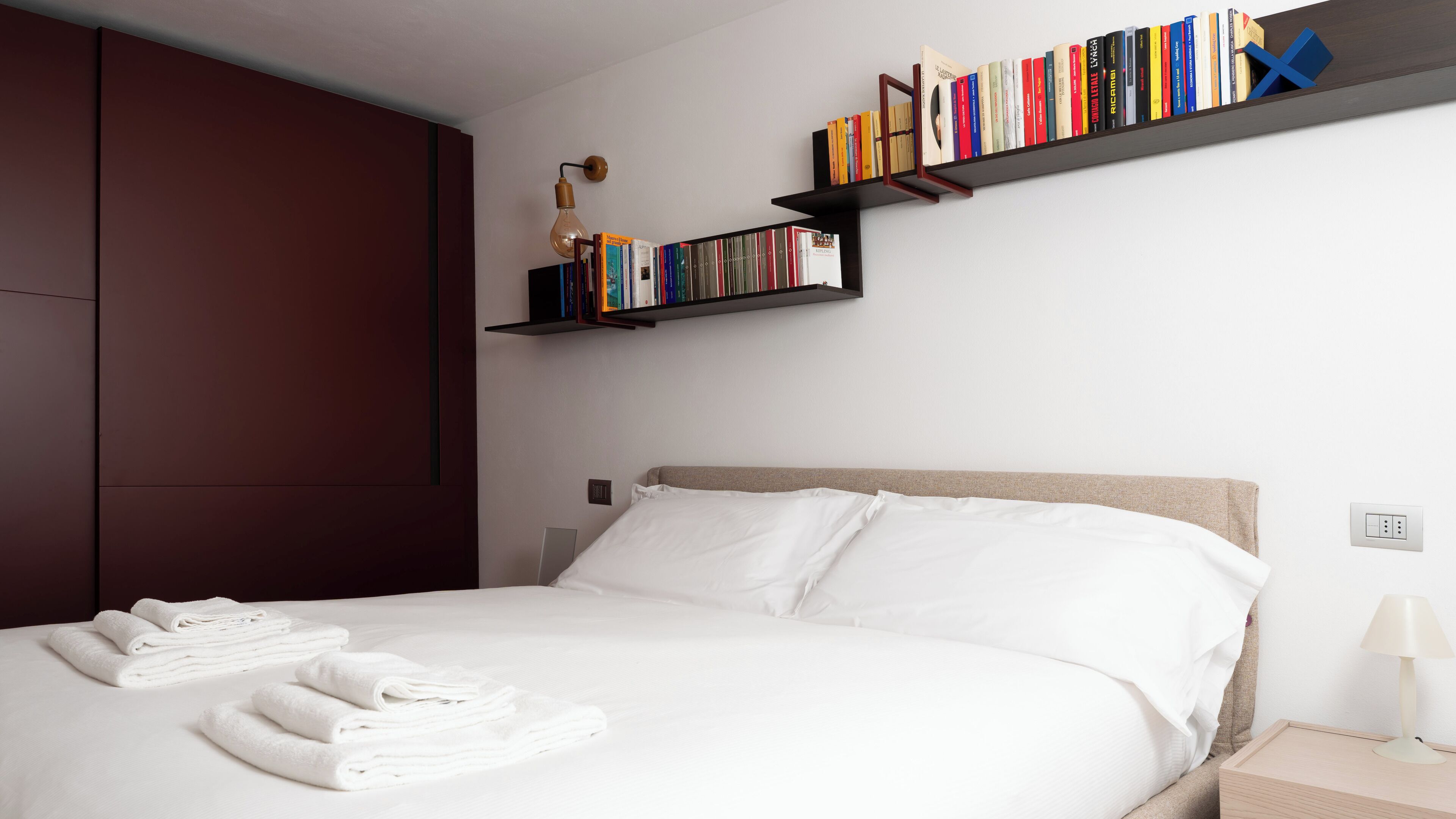 Una camera d'albergo con un letto, lenzuola bianche, un armadio in legno e due librerie piene di libri.