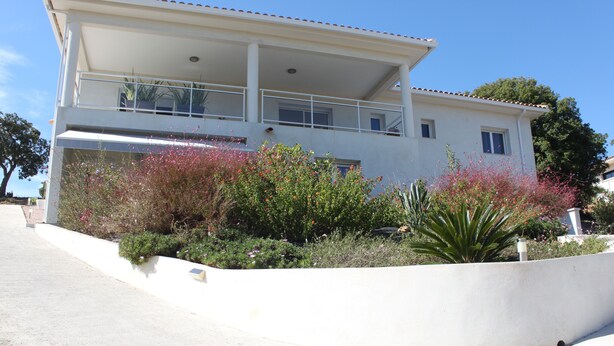 A two-story building with a balcony, surrounded by various plants and shrubs.
