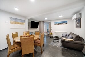 Un salon moderne avec une table à manger ronde, des chaises en osier, un canapé et un téléviseur à écran plat fixé au mur.