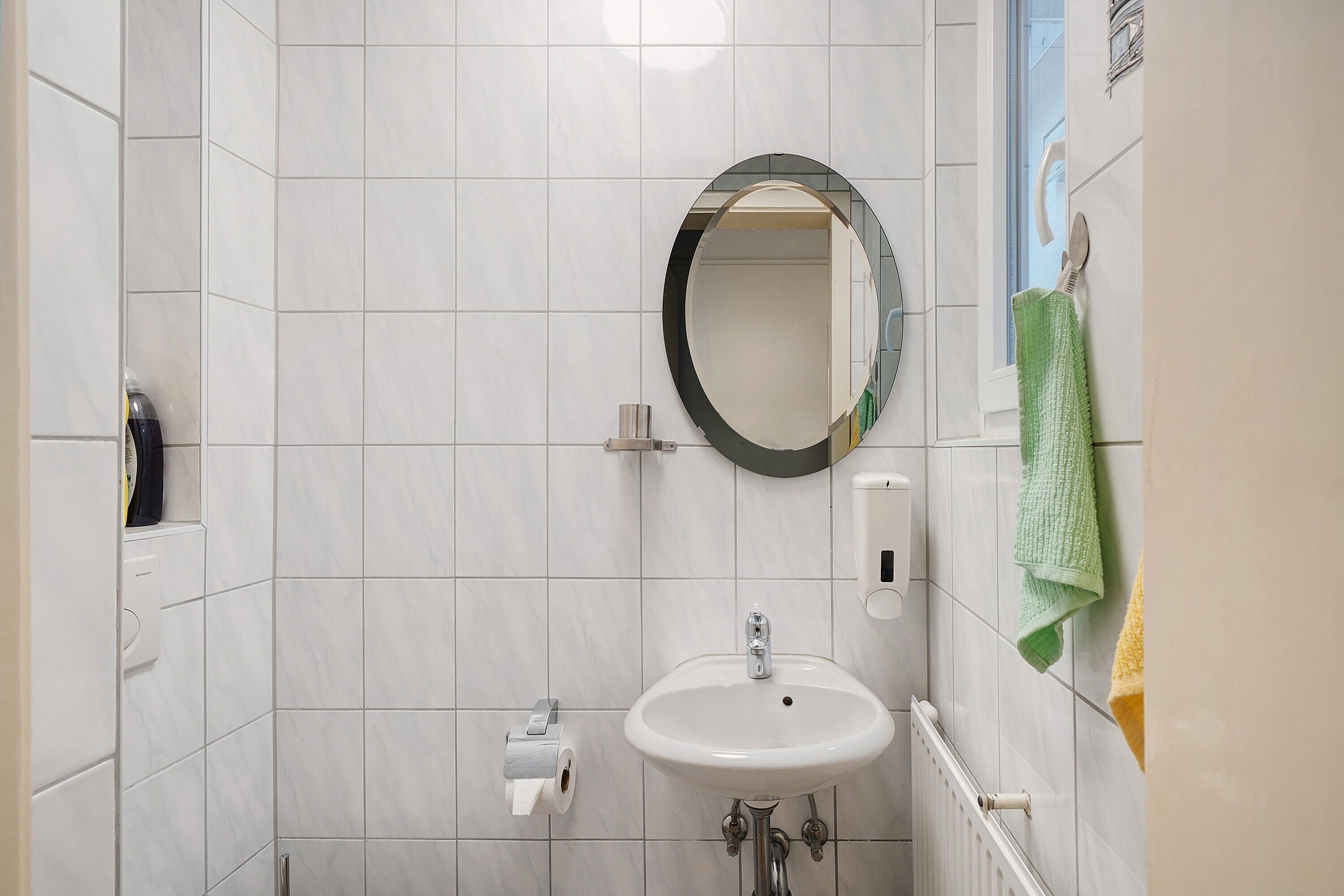 Ein Badezimmer mit einem weißen Waschbecken, einem Spiegel, einem Handtuchhalter mit grünen und gelben Handtüchern und einem Toilettenpapierhalter.