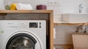 Een Bauknecht wasmachine in een moderne badkamer, met een houten plank erboven, een wastafel en een spiegel.