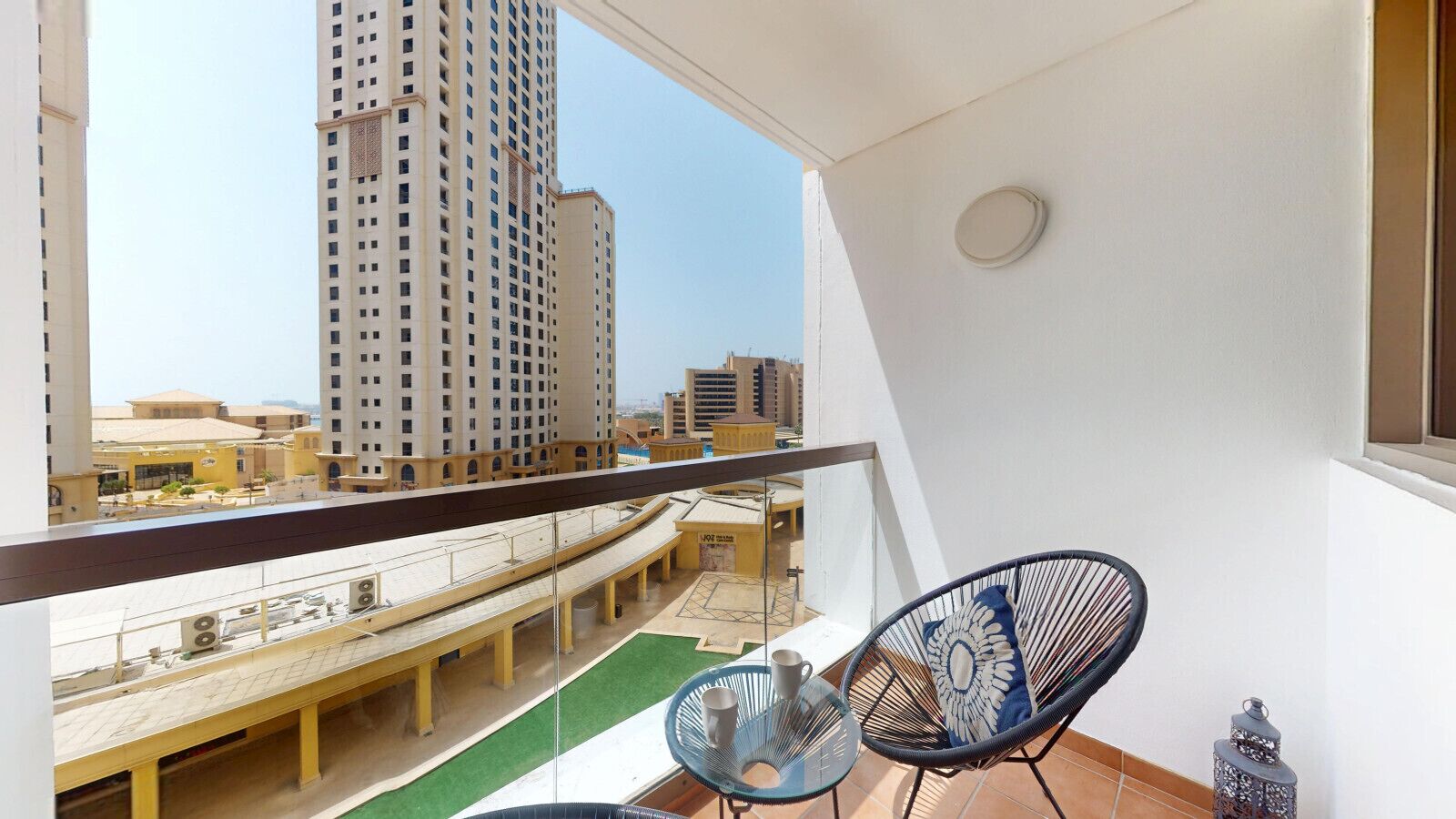 Un balcón con vistas a un paisaje urbano, que incluye edificios altos y un puente. Hay dos sillas y una mesita con un ventilador.