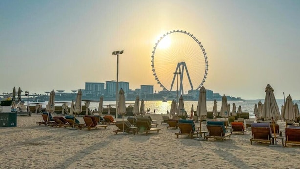 A beach with sun loungers and umbrellas, a Ferris wheel in the background, and a cityscape across the water.