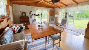 Een ruime woonkamer met een houten eettafel, een bank met kleurrijke kussens en een groot raam met uitzicht op de omgeving.