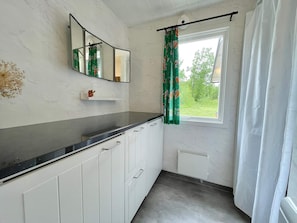Une salle de bain avec un plan de toilette noir, des meubles blancs, un miroir, une fenêtre avec un rideau et un radiateur.