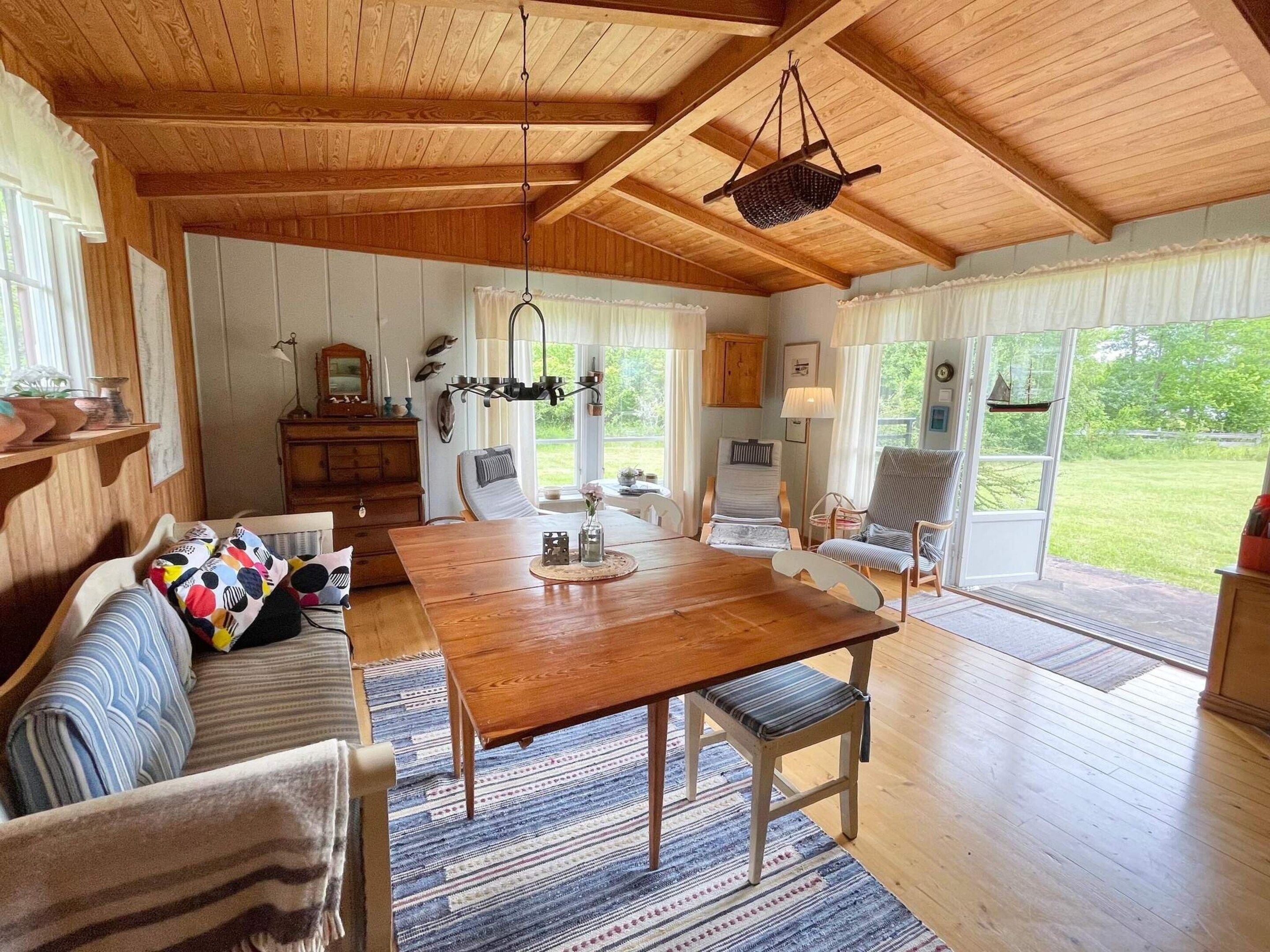 A cozy interior with a wooden dining table, a sofa with colorful pillows, and a view of the outdoors through large windows.