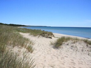 Une plage de sable avec quelques touffes d’herbe, un ciel dégagé et une mer calme.