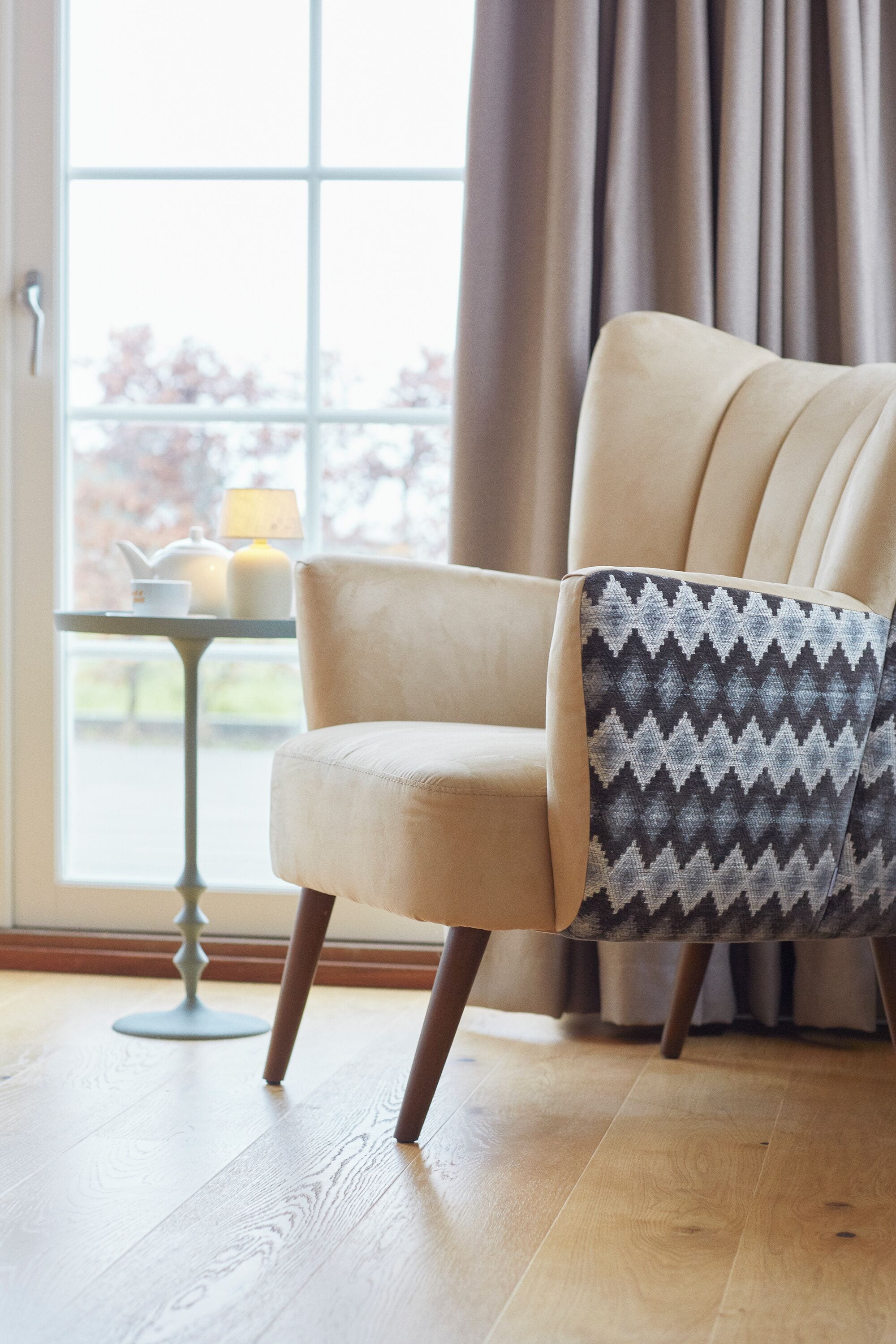Una silla con cojín estampado, una mesa con lámpara y una ventana con cortinas.