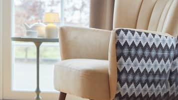 Una silla con cojín estampado, una mesa con lámpara y una ventana con cortinas.