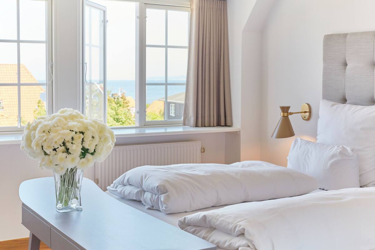 Ein Schlafzimmer mit einem Bett, einem Nachttisch mit Blumenvase, einem Fenster mit Meerblick und einer Wandlampe.