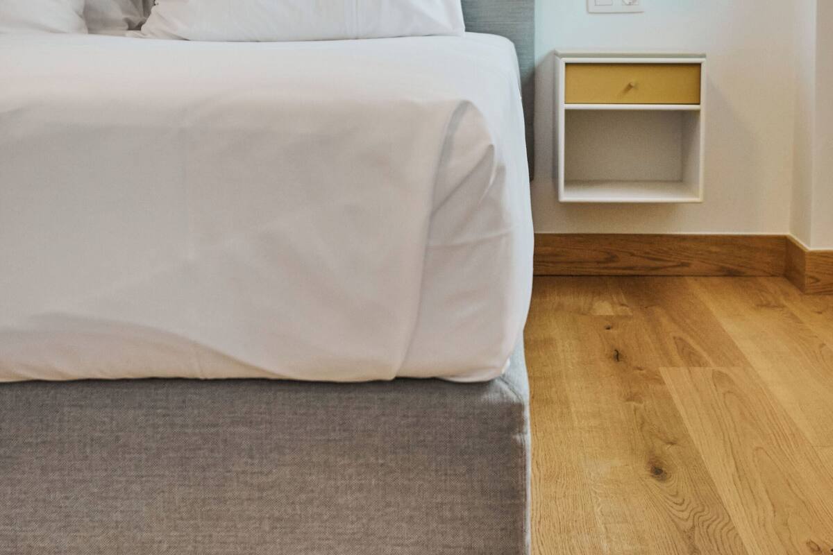 Ein ordentlich bezogenes Bett mit weißen Leinen, ein grau gepolsterter Kopfteil und ein Nachttisch mit einer Lampe.