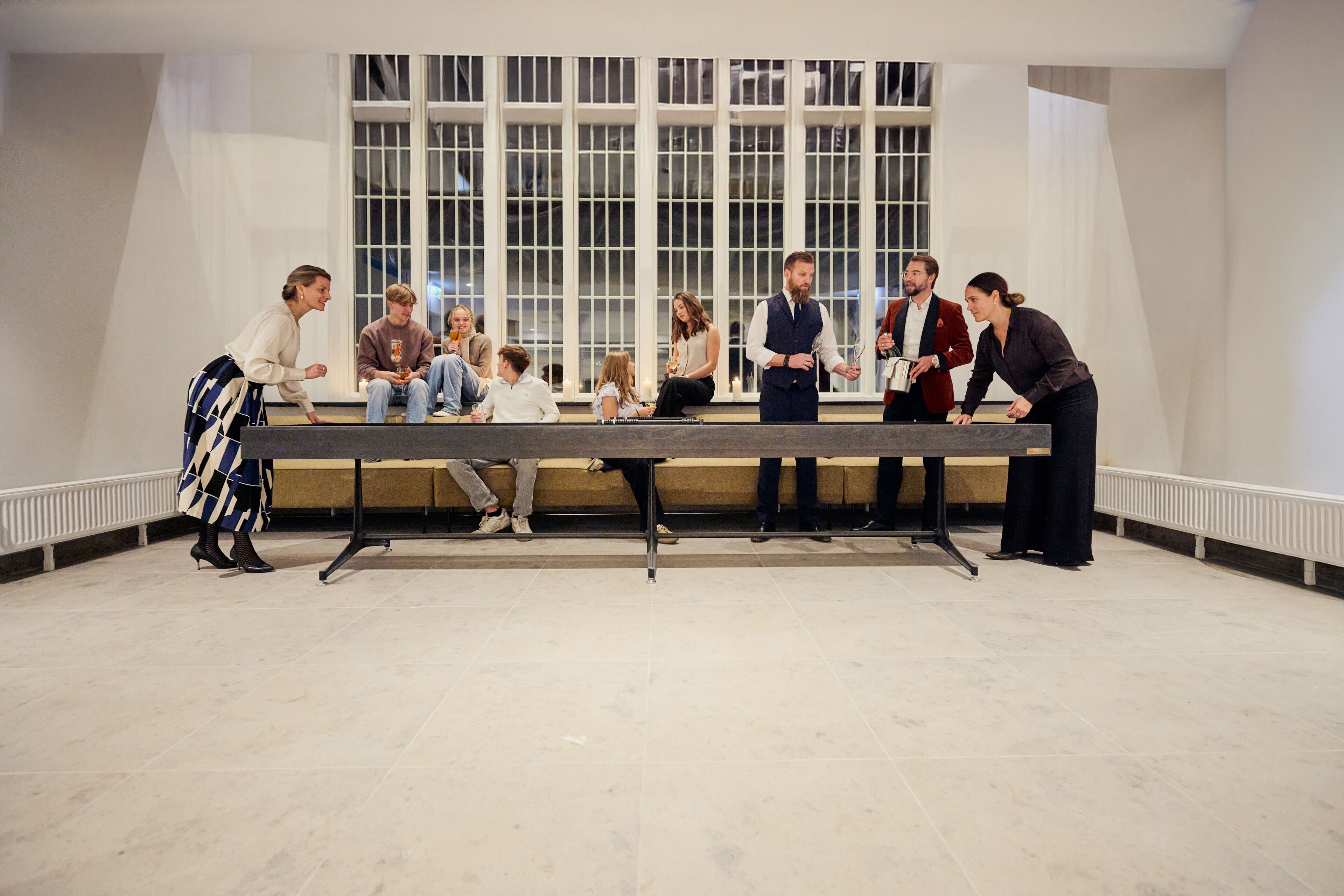 A group of people seated around a long table in a spacious room with large windows and a tiled floor.