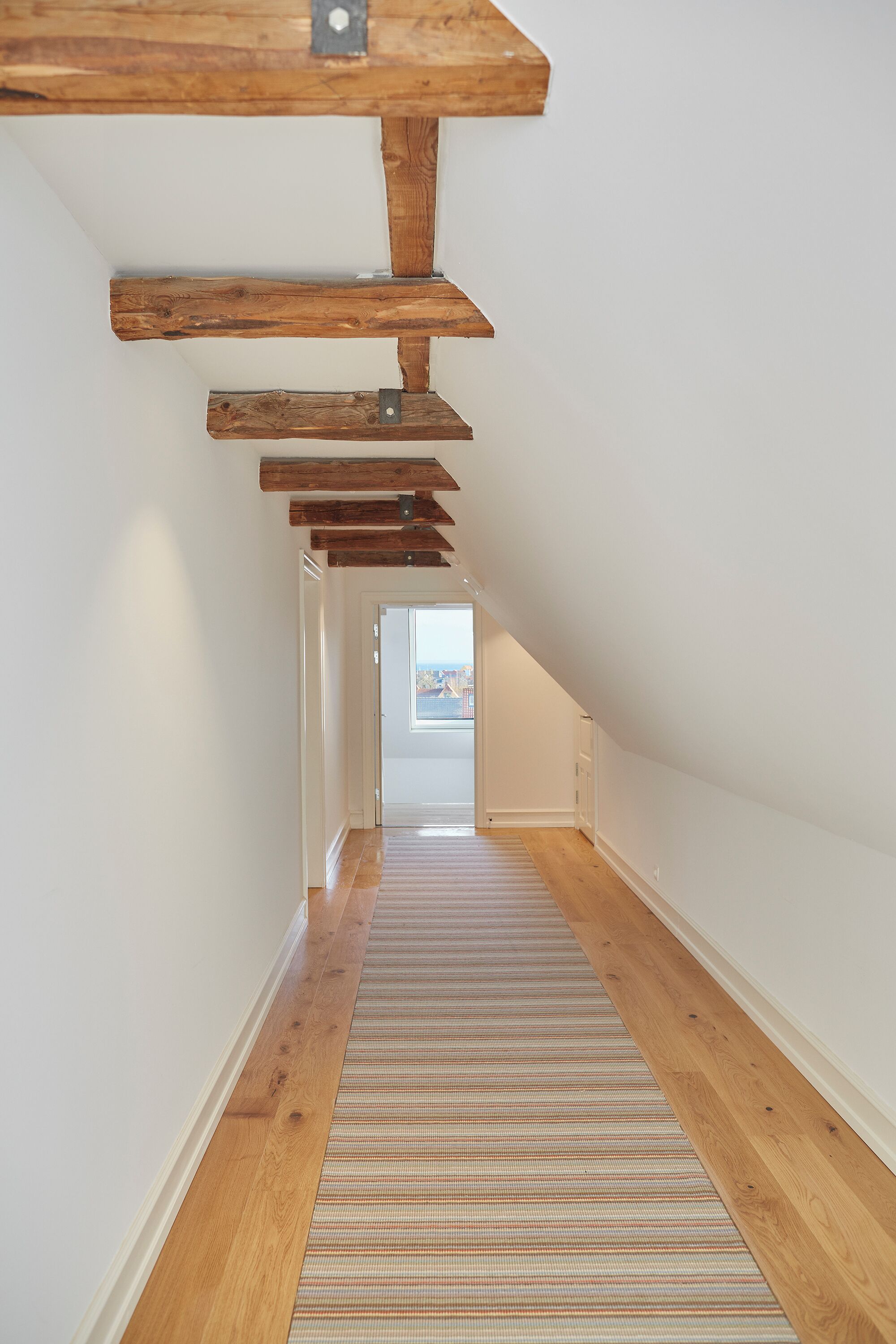 Un pasillo estrecho con vigas de madera, un suelo a rayas y vistas de la ciudad a través de una puerta al final.