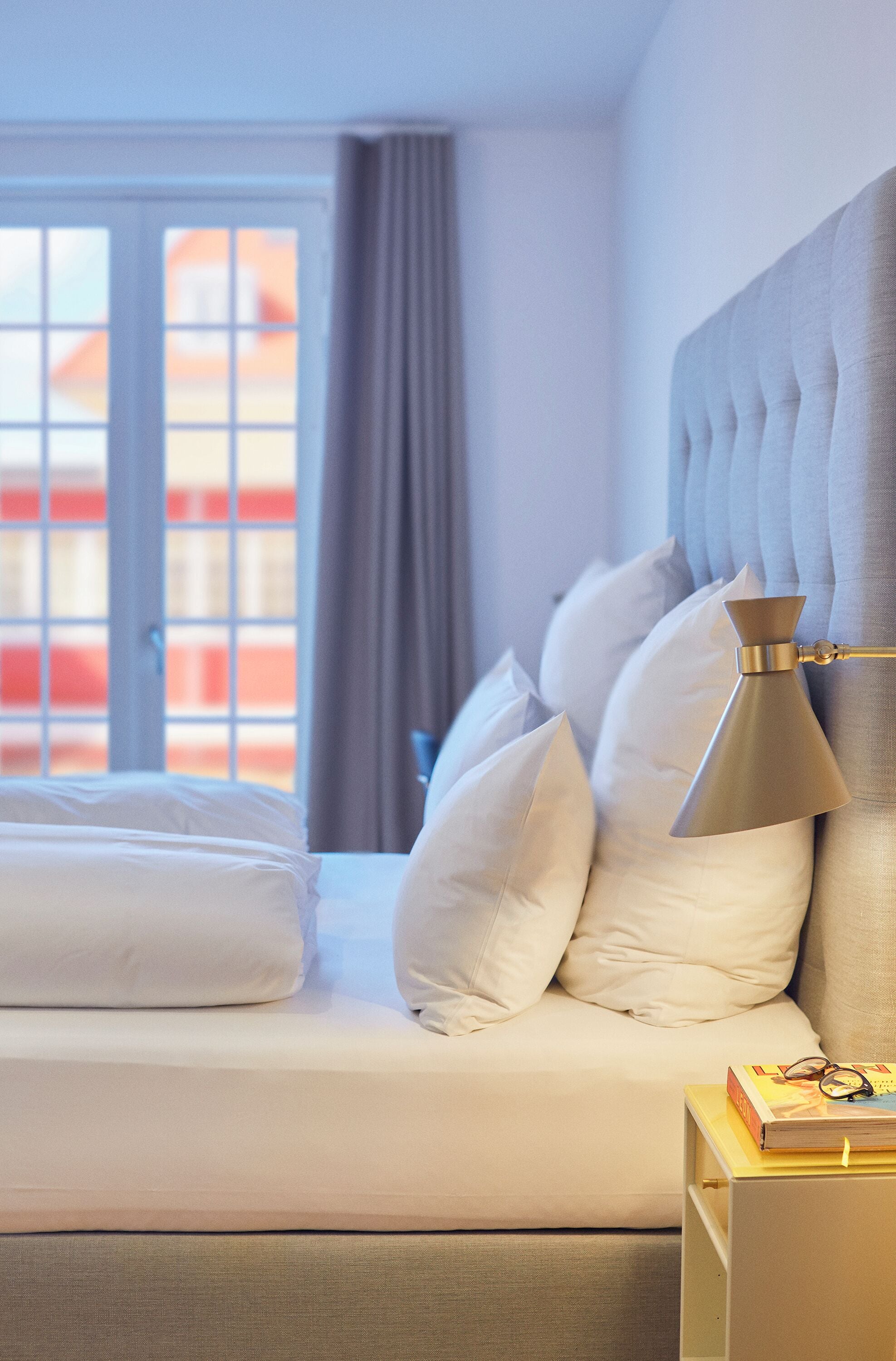 A neatly made bed with white linens and pillows, a bedside lamp, and a small table with books and glasses.