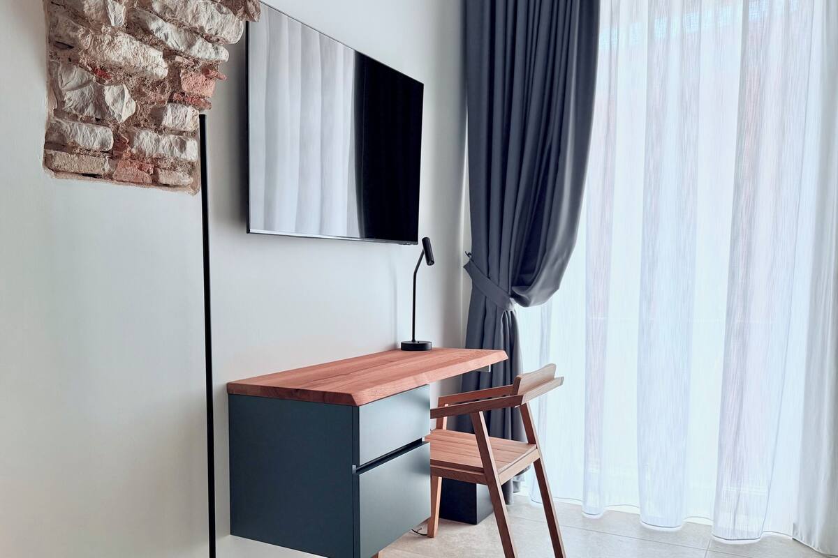 Een moderne werkruimte met een houten bureau, een stoel, een flatscreen televisie en een groot raam met dunne gordijnen.