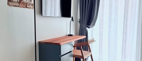 A modern workspace with a wooden desk, a chair, a flat-screen TV, and a large window with sheer curtains.