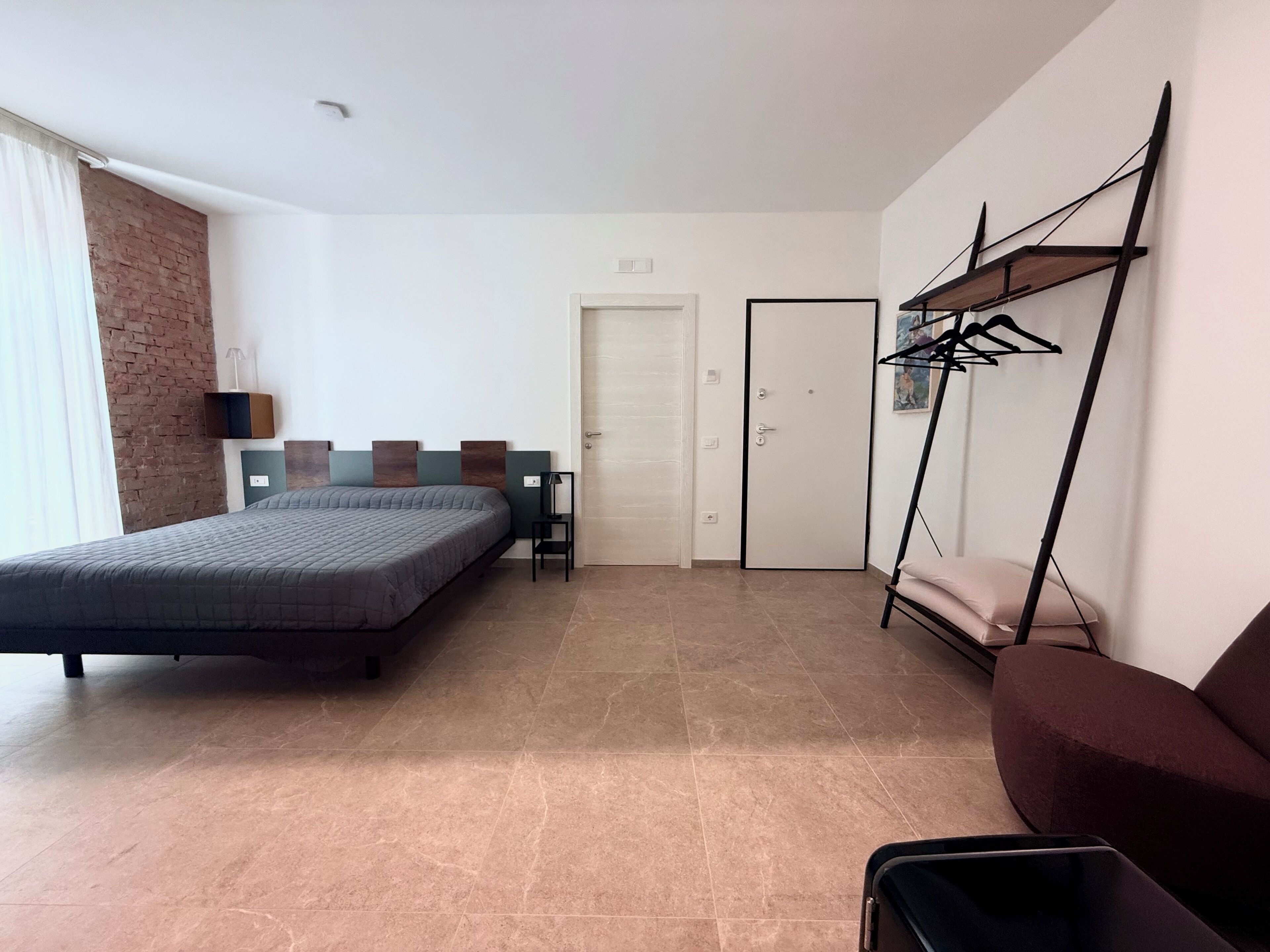 A modern hotel room with a large bed, a seating area with a chair and table, a wardrobe, and a hanging rack with clothes.