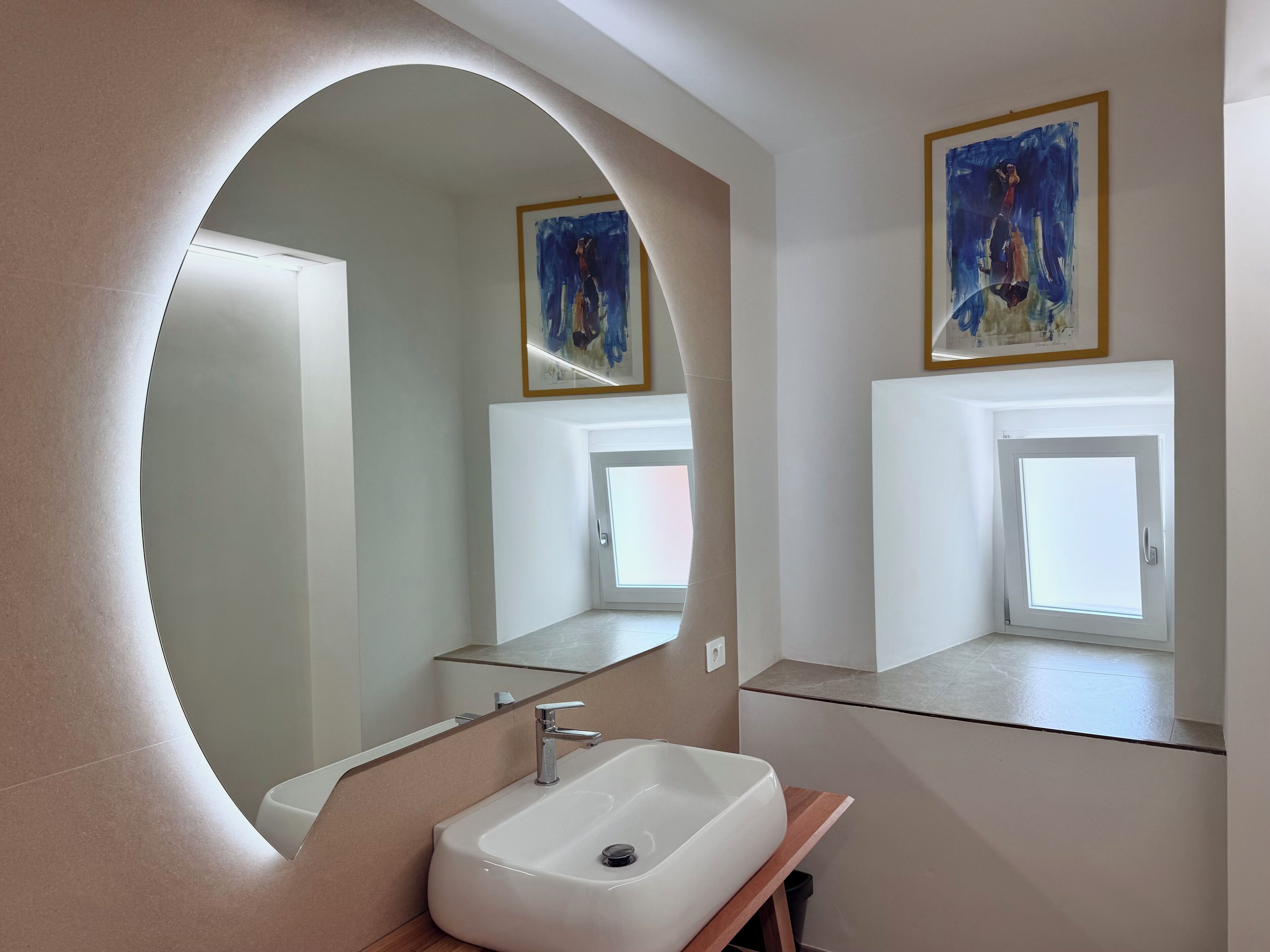 A modern bathroom with a large mirror, a white sink, and a window above the sink.