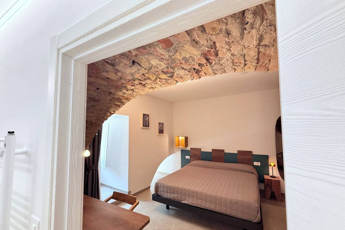 Een slaapkamer met een stenen plafond, een bed met een gebogen deken, een nachtkastje en een lamp.