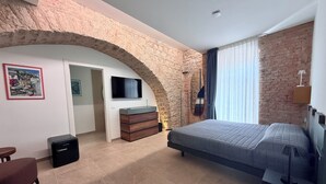 Ein Schlafzimmer mit einem Steingewölbe, einem Bett, einer Kommode, einem Fernseher und einem Fenster mit Vorhängen.
