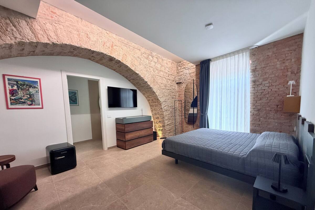 Een slaapkamer met een stenen boog, een bed, een dressoir, een televisie en een raam met gordijnen.